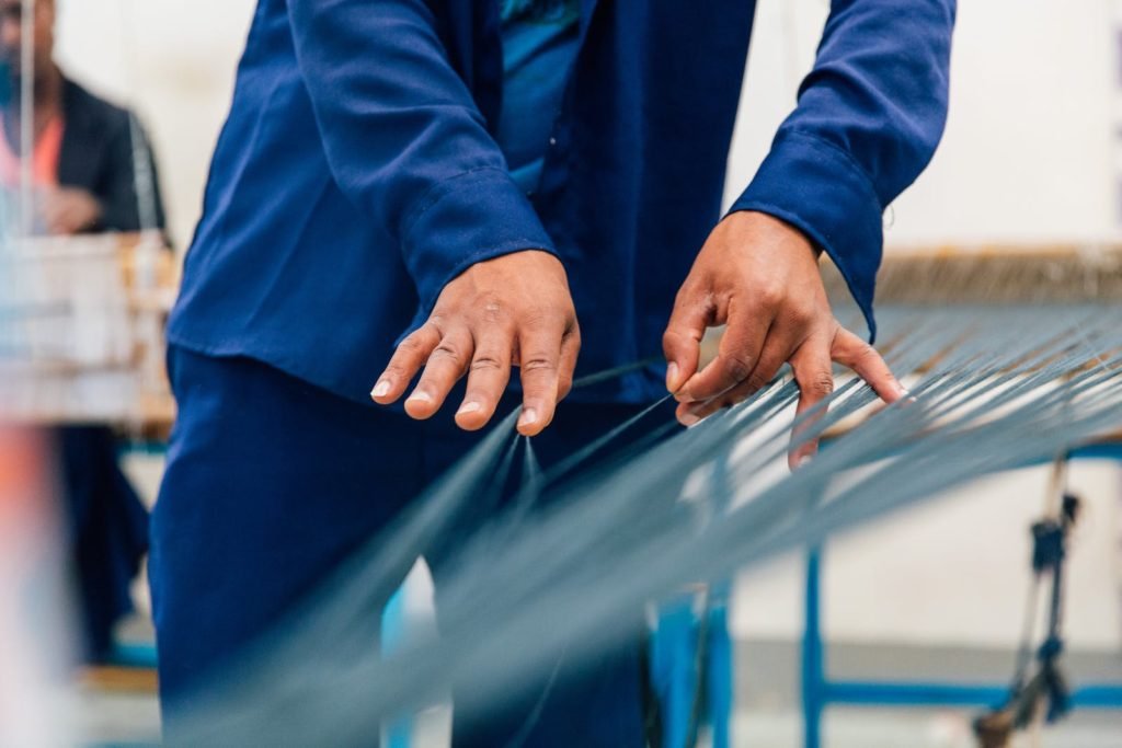 Crop unrecognizable craftsman in blue uniform unraveling thin threads on loom machine while working in studio with ethnic colleague